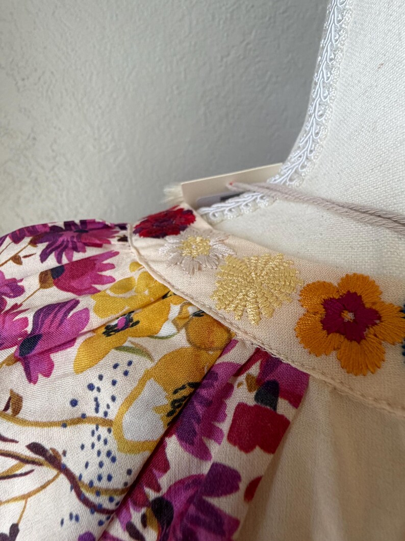 May include: Close-up of a cream-colored garment with floral print sleeves and embroidered details. The sleeves feature a vibrant pattern of purple, yellow, and pink flowers. The neckline is adorned with embroidered yellow and orange flowers.