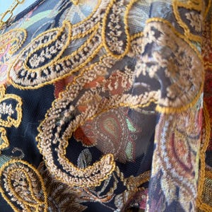 May include: Close-up of a sheer fabric with intricate embroidery. The design features paisley patterns in shades of brown, orange, and green, outlined with gold thread. A gold chain is visible in the upper left corner.