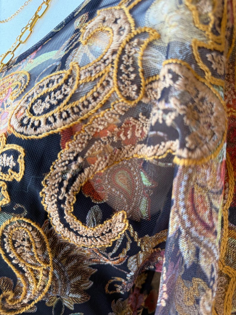 May include: Close-up of a sheer fabric with intricate embroidery. The design features paisley patterns in shades of brown, orange, and green, outlined with gold thread. A gold chain is visible in the upper left corner.