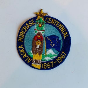 May include: A blue and yellow embroidered patch with the text "Alaska Purchase Centennial 1867-1967". The patch features a cartoon image of a totem pole with a star on top.