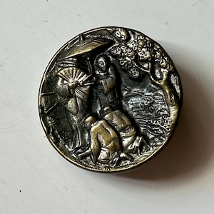 May include: A round, metallic button with a dark, antiqued brass finish. The surface features a raised, detailed relief carving depicting figures in traditional attire, one holding an umbrella, alongside a tree and landscape elements.