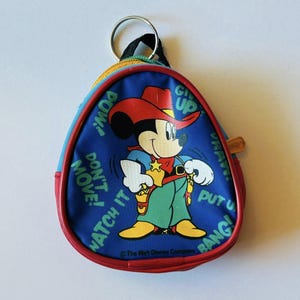 May include: A blue, red, and yellow Mickey Mouse coin purse with a cowboy theme. The purse features Mickey Mouse in a cowboy hat and green pants, with text that reads "POW!", "DON'T MOVE!", "WATCH IT", "GIVE UP", and "PUT 'EM UP BANG!"