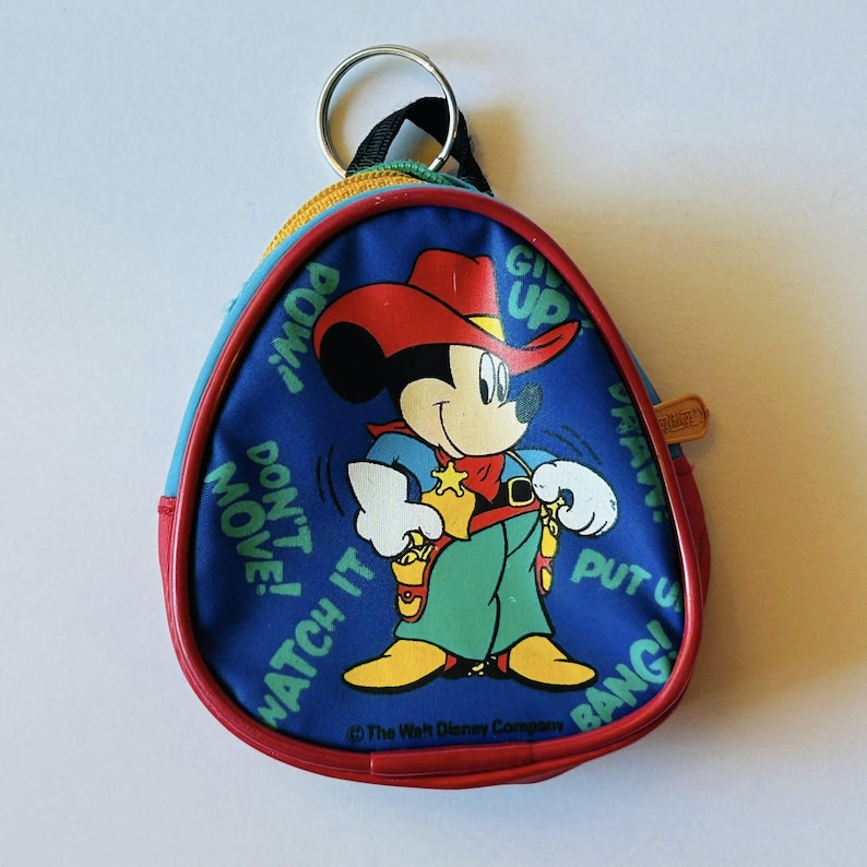 May include: A blue, red, and yellow Mickey Mouse coin purse with a cowboy theme. The purse features Mickey Mouse in a cowboy hat and green pants, with text that reads "POW!", "DON'T MOVE!", "WATCH IT", "GIVE UP", and "PUT 'EM UP BANG!"