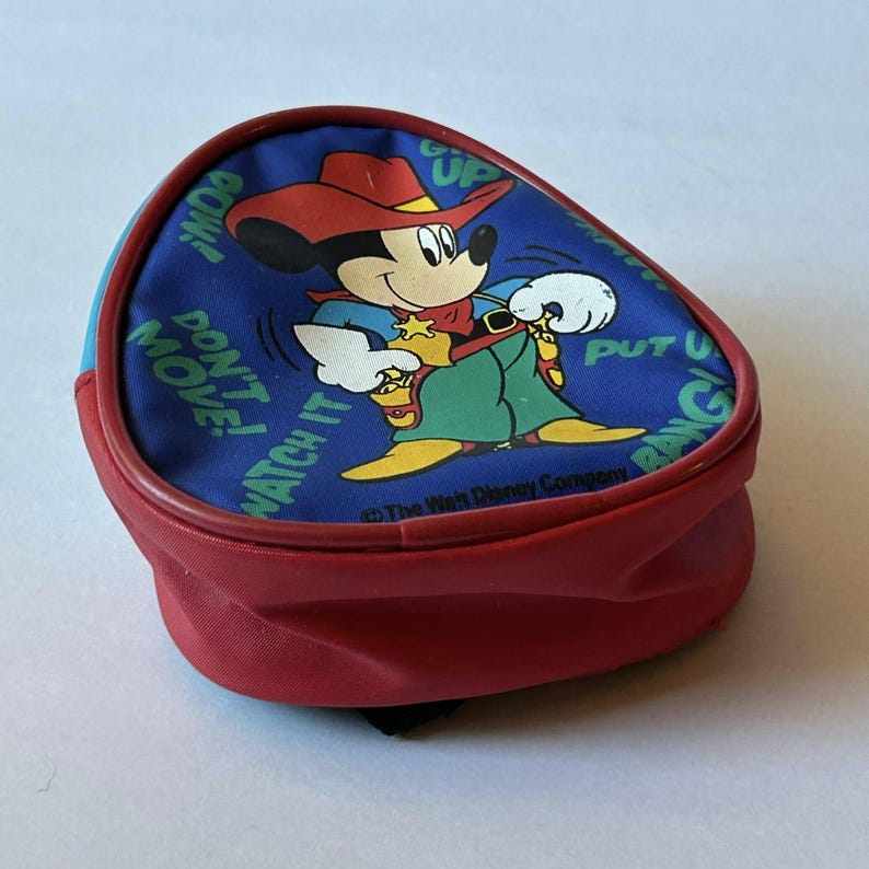 May include: A small, red backpack with a blue top featuring a cartoon Mickey Mouse dressed as a cowboy. The design includes the phrases "Watch it", "Don't Move", and "Put 'em up". The backpack has a black strap.