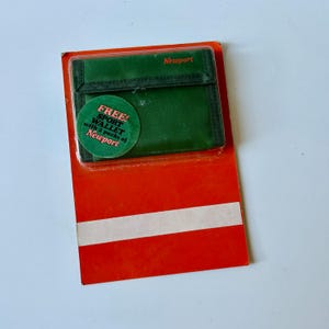 May include: A green fabric tri-fold sport wallet with the word Newport printed in orange on the flap. The wallet is encased in clear plastic on a bright orange cardboard backing featuring a circular sticker that reads FREE! SPORT WALLET with 2 packs of Newport.
