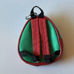 May include: A miniature backpack keychain with a green body, red straps, and a red border. The small backpack has black straps and a silver metal ring for attaching to keys or a bag. The backpack is a fun accessory.