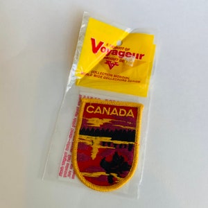 May include: A Canada patch with a yellow border, featuring the word "CANADA" in red letters. The patch depicts a landscape scene with a canoe, trees, and a sunset. The patch is in a clear plastic bag with a yellow tag that reads "A PRODUCT OF Voyageur".