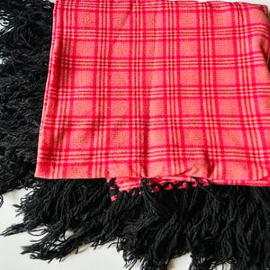 May include: A red and pink plaid scarf with black fringe. The scarf is folded, displaying the plaid pattern prominently. The black fringe is thick and textured, creating a contrast with the smoother plaid fabric. This scarf is a fashion accessory.