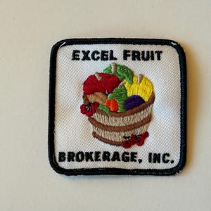 May include: White embroidered patch with black border. The patch features a basket of fruit and the text "EXCEL FRUIT BROKERAGE, INC."