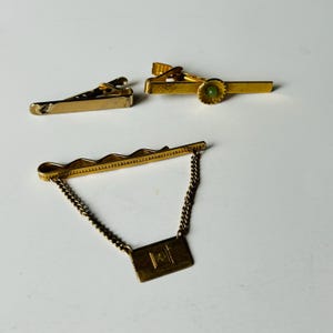 May include: Three gold-toned tie clips are displayed against a white background. One clip has a textured surface, another features a green stone, and the third has a chain and a rectangular emblem. These vintage accessories are suitable for formal wear.