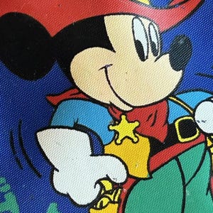 May include: Close-up of a colorful cartoon image of Mickey Mouse dressed as a cowboy. Mickey is wearing a red hat, blue shirt, red cape, and green pants. A yellow star-shaped badge is on his chest. The background is blue.