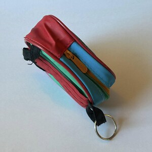 May include: A small, rectangular, multi-colored zippered pouch. The top is red, with blue, green, and yellow sections. It has a gold zipper and a black strap with a silver key ring attached. The pouch is likely for carrying small items.