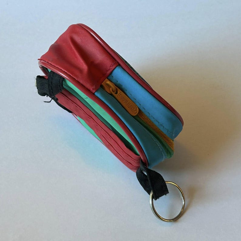 May include: A small, rectangular, multi-colored zippered pouch. The top is red, with blue, green, and yellow sections. It has a gold zipper and a black strap with a silver key ring attached. The pouch is likely for carrying small items.