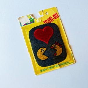 May include: Iron-on patch featuring a red heart and two Pac-Man-like characters in yellow and red, on a dark blue denim background. The patch is packaged in a yellow cardboard backing with the words "IRON-ON" and "QUICK EASY ECONOMICAL".