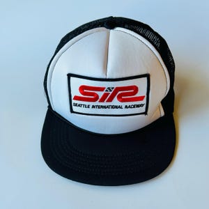 May include: A black and white trucker hat with a white front panel and a black mesh back. The hat features a rectangular patch with the red text "SIR" and "SEATTLE INTERNATIONAL RACEWAY". The brim is black.