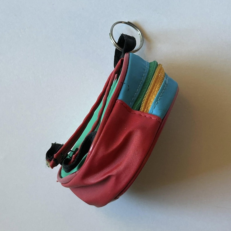 May include: A miniature, colorful backpack keychain. The keychain features a red base, blue top, and green and yellow zippers. The small backpack has a black strap and a silver key ring.