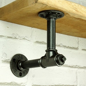 Puede incluir: Soporte de estante de tubería de metal negro con un estante de madera. El soporte está fijado a una pared de ladrillo blanco. El soporte tiene un diseño en forma de T con una brida redonda en cada extremo. La brida del lado izquierdo tiene los números "DS" y "SO" impresos en ella. La brida del lado derecho tiene el número "33" impreso en ella.