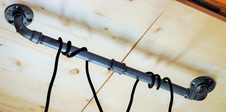 May include: A black metal pipe with two flanges on either end, mounted to a wooden ceiling. Two black cords are hanging from the pipe.