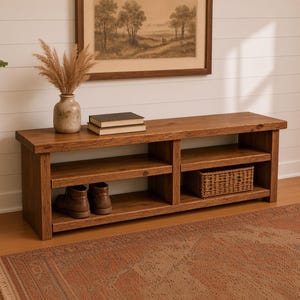 May include: A rectangular wooden media console featuring two open shelves, a smooth top surface, and a sturdy base. The console is constructed from medium-toned wood with visible grain patterns, providing storage space for books, a woven basket, and footwear.