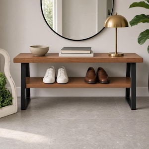 May include: A rectangular wooden bench with two tiers and black metal legs. The top surface holds a beige bowl, two stacked books, and a gold dome-shaped lamp. The lower shelf displays a pair of white sneakers and a pair of brown leather loafers.