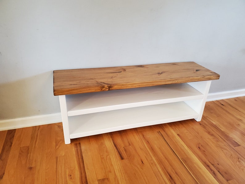May include: A white wooden bench with a natural wood top and two shelves. The bench is sitting on a hardwood floor.