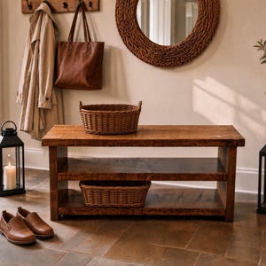 May include: A rectangular wooden bench with two open shelves, featuring a dark brown finish. A woven wicker basket sits on the top surface and another on the bottom shelf. The bench is positioned on a stone floor between two black metal lanterns.