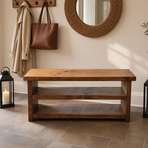 May include: A rectangular wooden bench featuring a natural brown finish with a smooth top surface and a single open shelf underneath, constructed with thick, sturdy planks and visible wood grain patterns throughout the entire furniture piece.
