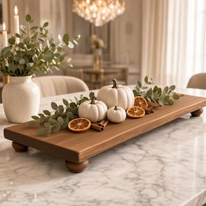 May include: A rectangular wooden riser with four rounded feet, topped with three white pumpkins, dried orange slices, cinnamon sticks, and green eucalyptus sprigs. The tray features a smooth, medium-brown wood grain finish and a flat surface.