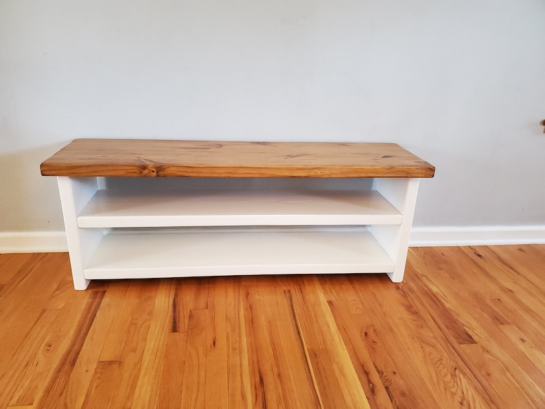 May include: A white wooden bench with a natural wood top and two shelves. The bench is sitting on a hardwood floor.