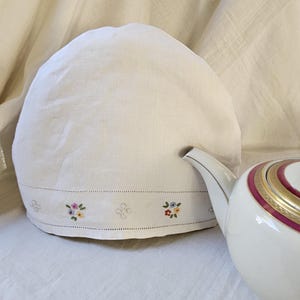 May include: A white tea cozy with a floral embroidered border, next to a white teapot with a gold and burgundy band. The cozy is made of a soft, textured fabric.