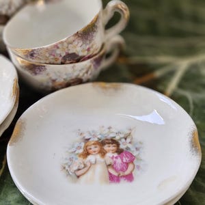 May include: A stack of white porcelain teacups with gold trim and floral patterns. A matching saucer with a painted image of two girls in dresses and gold trim. The teacups and saucers are part of a vintage tea set.