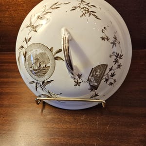 May include: A white ceramic lid with a brown floral design and a handle. The lid has a circular design with a ship in the center.