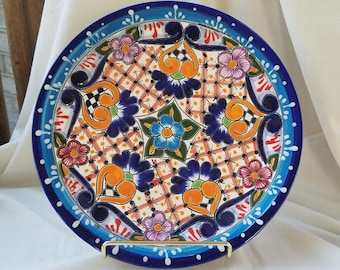 May include: A large, round ceramic plate with a colourful Mexican Talavera design. The plate features a central motif of a blue flower with orange, green, and yellow accents. The border of the plate is decorated with a blue and white pattern, and the plate is accented with white dots.