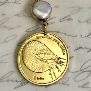 May include: Gold-toned necklace featuring a coin pendant with a nautilus design and the text "Nautilus pompilius". The necklace has a pearl accent and a toggle clasp. The chain is made of oval links.