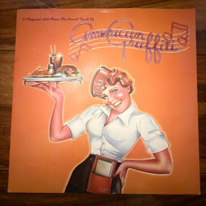 May include: Vintage album cover for "American Graffiti" soundtrack. The cover features a retro illustration of a smiling waitress holding a tray of food. The title is in a neon-style font, set against an orange background.