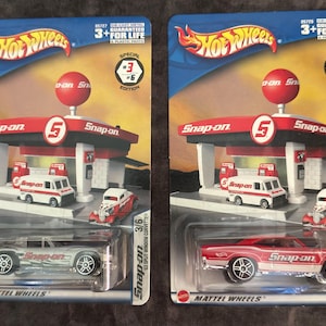 May include: Two Hot Wheels die-cast car sets featuring Snap-on gas station models and matching cars. One set includes a silver '63 Split Window Corvette, the other a red Road Runner. Both sets have the Hot Wheels logo and "3+ FOR LIFE" text.
