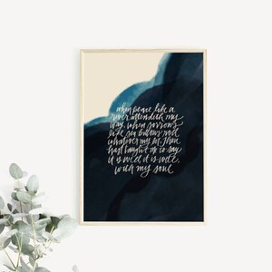 May include: A rectangular wall art print in a light wood frame featuring a dark navy blue and cream watercolor background. The print displays white cursive calligraphy text that reads: When peace like a river, attendeth my way, when sorrows like sea billows roll; whatever my lot, Thou hast taught me to say, it is well, it is well, with my soul.
