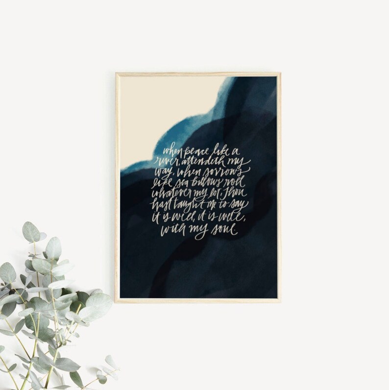 May include: A rectangular wall art print in a light wood frame featuring a dark navy blue and cream watercolor background. The print displays white cursive calligraphy text that reads: When peace like a river, attendeth my way, when sorrows like sea billows roll; whatever my lot, Thou hast taught me to say, it is well, it is well, with my soul.