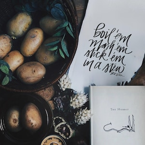 Op de afbeelding: Een rustig stilleven met aardappelen in een rieten mand en een donkere kom. Een handgeschreven citaat van J.R.R. Tolkien luidt "Boil 'em, mash 'em, stick 'em in a stew." Een exemplaar van "The Hobbit" is ook aanwezig.