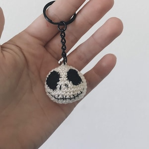May include: A crocheted keychain with a round, off-white ball and a black Jack Skellington face. The keychain has a black metal ring and chain. The face has black stitched eyes and a stitched mouth. The keychain is held in a hand.