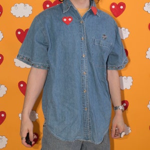 May include: A light blue denim shirt with short sleeves and a button-up front. The shirt has a small red heart with eyes on the left side of the chest and a small embroidered design on the left pocket.
