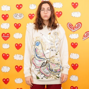 May include: A cream-colored sweater with a large embroidered butterfly design. The butterfly is made of silver, gold, pink, blue, and white thread. The sweater has long sleeves and a crew neck.