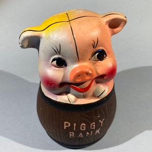 May include: A ceramic piggy bank shaped like a pig's head with a yellow and white face and a brown barrel body. The pig has a pink snout and a red mouth. The barrel has the words "Piggy Bank" written on it.
