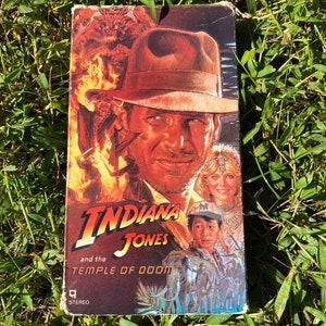 May include: A VHS tape cover for the movie Indiana Jones and the Temple of Doom. The cover features a portrait of Indiana Jones, a woman with a turban, and a young boy. The title of the movie is in large, red letters.