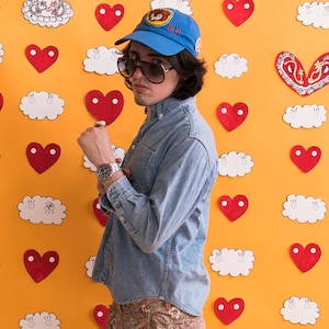 May include: A man wearing a blue denim shirt, a blue baseball cap with a patch that says "BLM", and sunglasses. He is standing in front of a yellow wall with red hearts and white clouds.