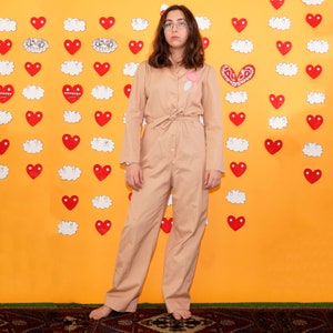 May include: A woman wearing a light brown jumpsuit with a pink heart design on the chest. She is standing in front of a yellow wall with red hearts and white clouds.