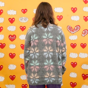 May include: A gray sweater with a pink, blue, and white snowflake pattern. The sweater is worn by a person standing in front of a yellow wall decorated with hearts and clouds.