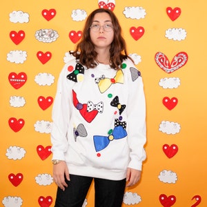May include: A white sweatshirt with colorful bows and buttons. The sweatshirt has a playful and whimsical design.