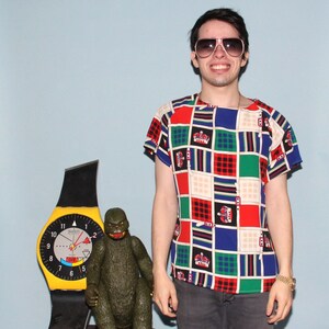 May include: A man wearing a colorful, geometric patterned short-sleeved shirt with a red, white, and blue color scheme. He is wearing sunglasses and has a smile on his face. A yellow and black Swatch watch is in the background.