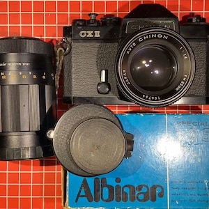 May include: A black Chinon CX II 35mm film camera with a 1:1.4 155mm Auto Chinon lens attached. The camera is sitting on a red grid surface with a black lens cap and a blue box with the text "SPECIAL 3" and "Albinar" printed on it.
